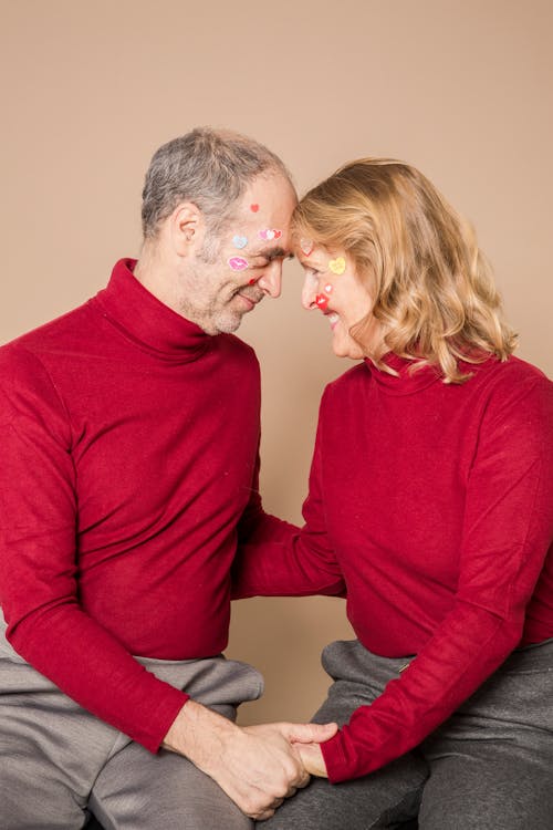 Beloved senior couple in stylish red turtlenecks with face paintings holding hands and touching foreheads while sitting against beige background in studio