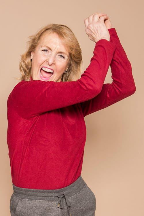 Joyful mature female with blond hair in red sweater yelling and looking at camera while dancing happily in beige studio
