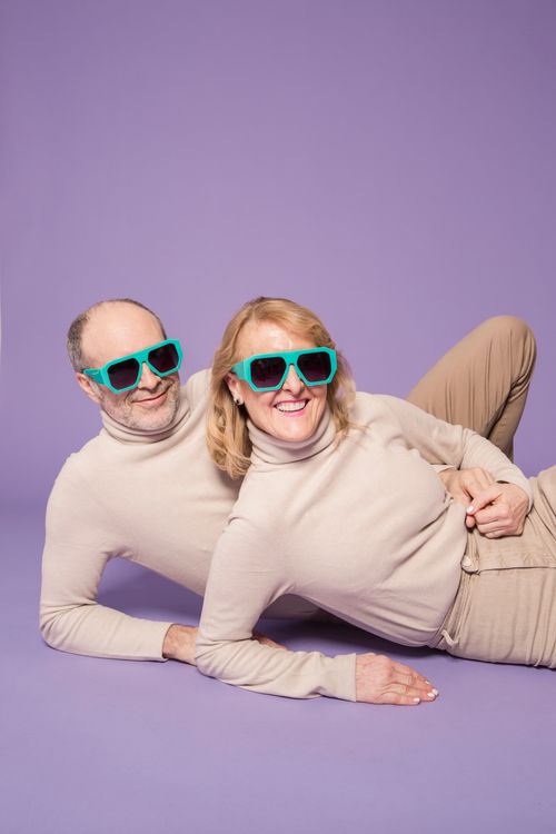 Free A joyful senior couple posing in matching outfits and sunglasses against a purple backdrop. Stock Photo