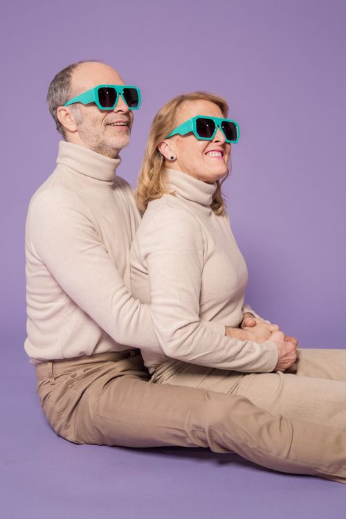 Free Elderly couple wearing matching sweaters and sunglasses, sitting with joy on a purple backdrop. Stock Photo