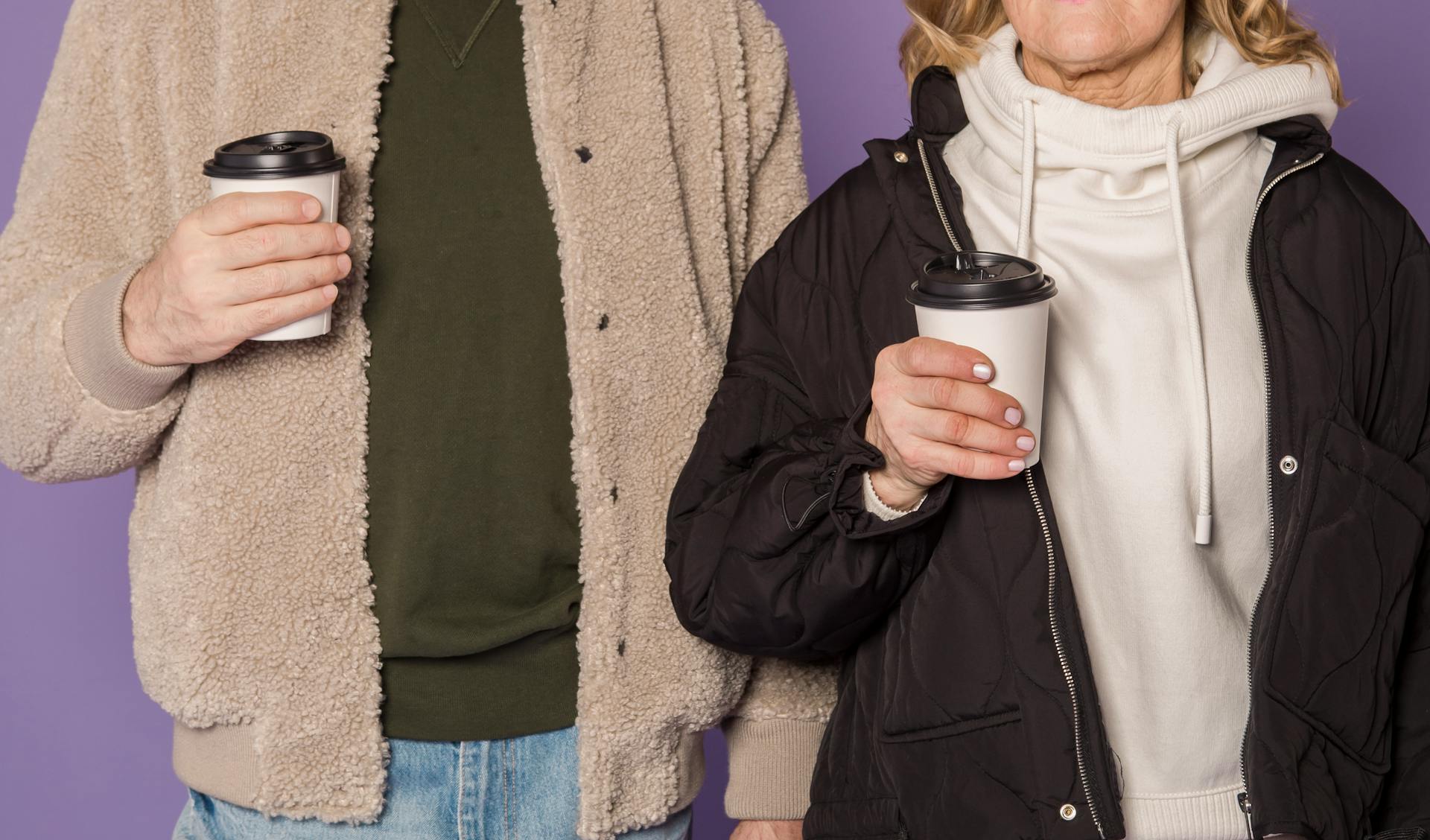 Senior couple in casual winter jackets enjoying coffee against a purple background.