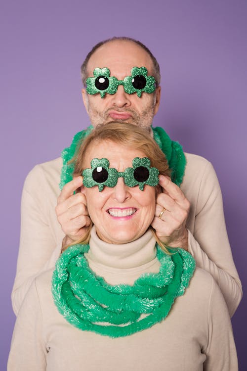 Cheerful middle aged couple wearing fancy party sunglasses and necklace while celebrating St Patricks Day