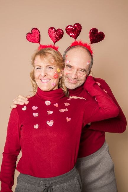 Fotomural Feliz pareja de ancianos celebrando el Día de San Valentín con accesorios en forma de corazón y suéteres rojos, abrazándose con cariño.