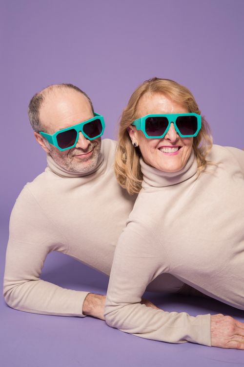 Free Happy senior couple posing playfully in matching sweaters and sunglasses against a purple background in a studio. Stock Photo