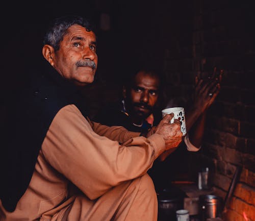 Old Indian man with gray hair and mustache sitting with cup of hot drink in brick poor building