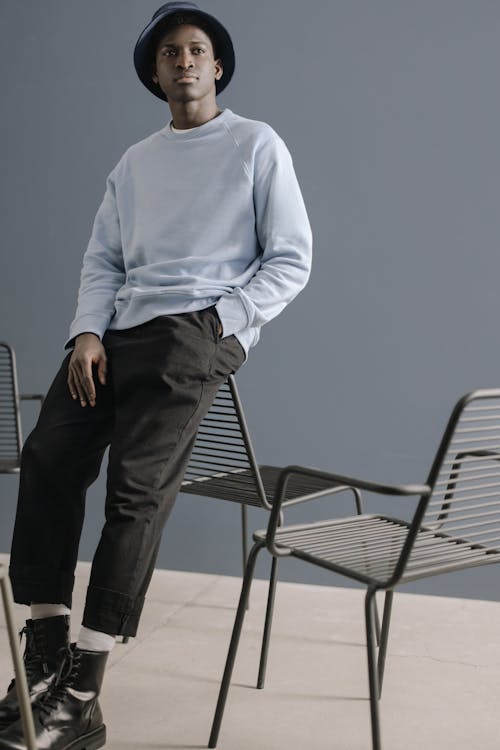 Man in Blue Sweater and Black Pants Leaning on Black Metal Chair 