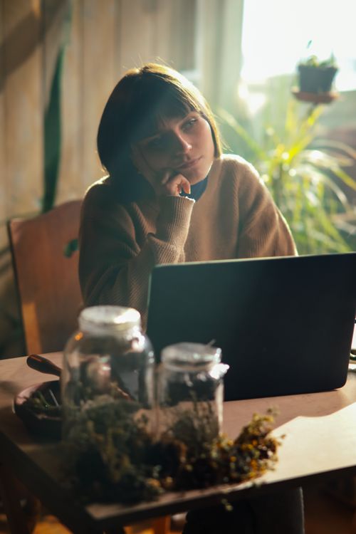 Free Thoughtful woman working on laptop surrounded by plants in a cozy indoor setting. Stock Photo