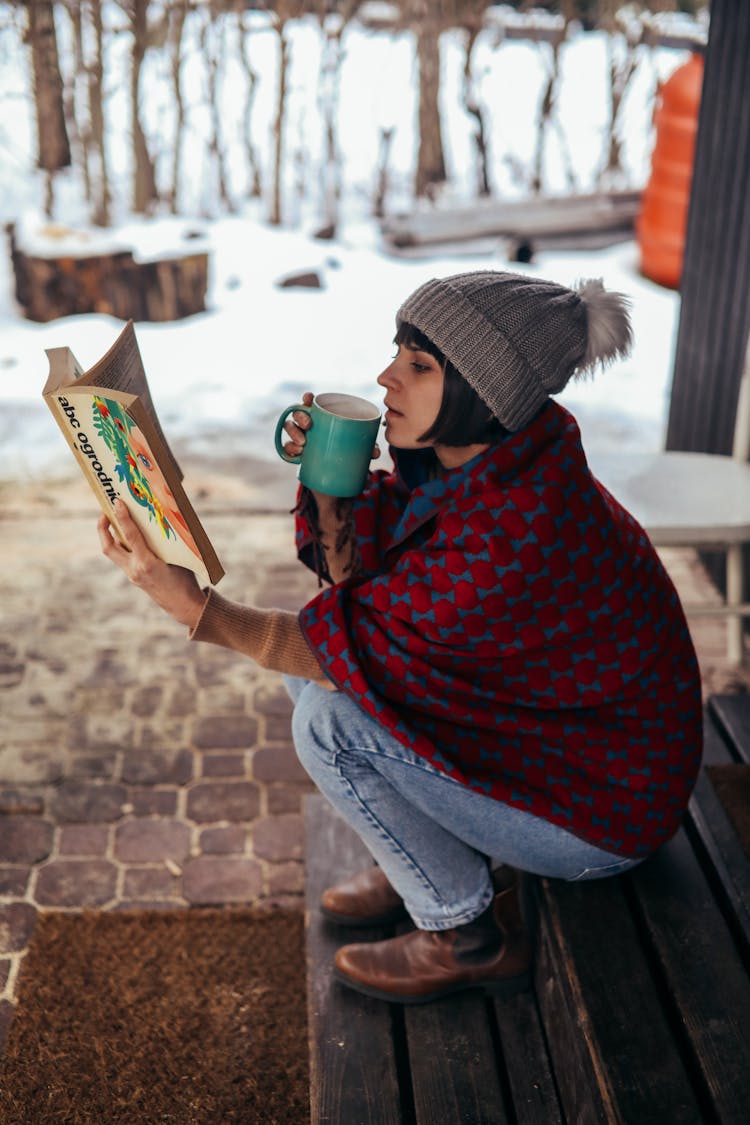 
A Woman Covered In A Blanket Reading A Book While Drinking Coffee