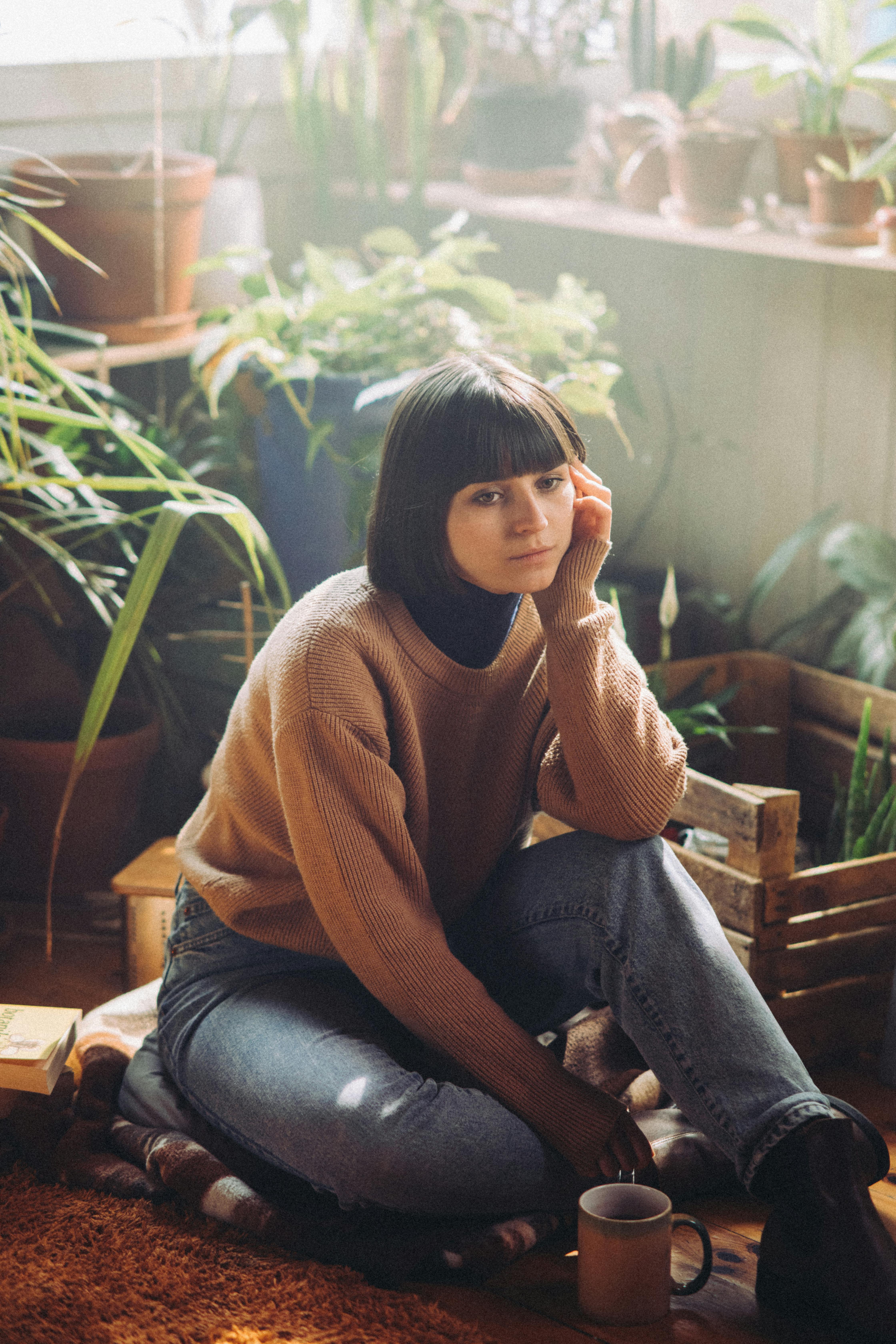 Woman sitting indoors surrounded by plants, wearing denim and sweater.