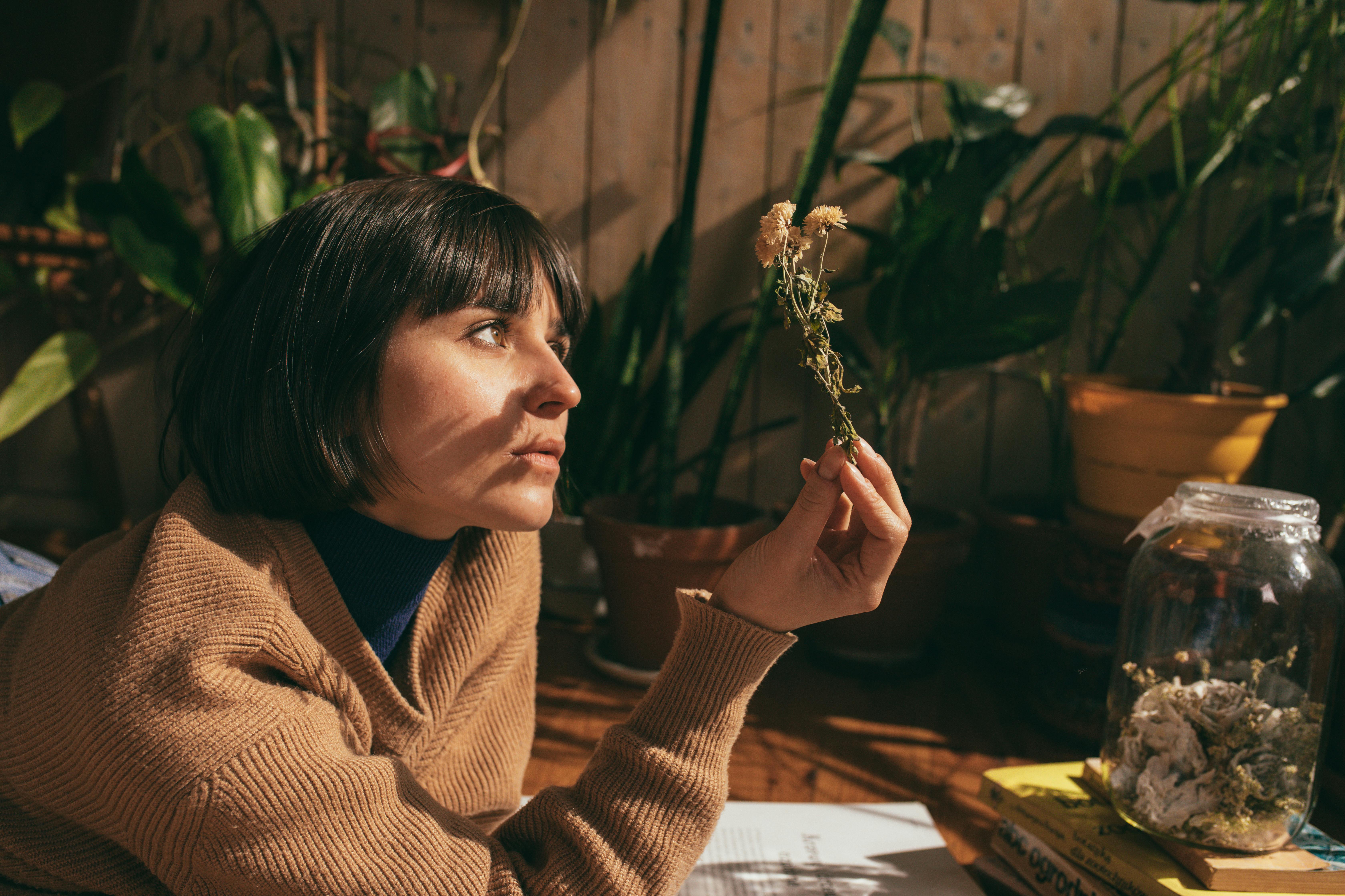 A woman with short hair in a sweater holds dried flowers indoors, surrounded by plants.