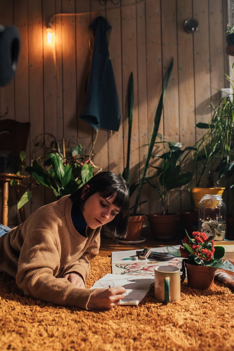 A Woman In A Sweater Lying Down On A Carpet While Drawing