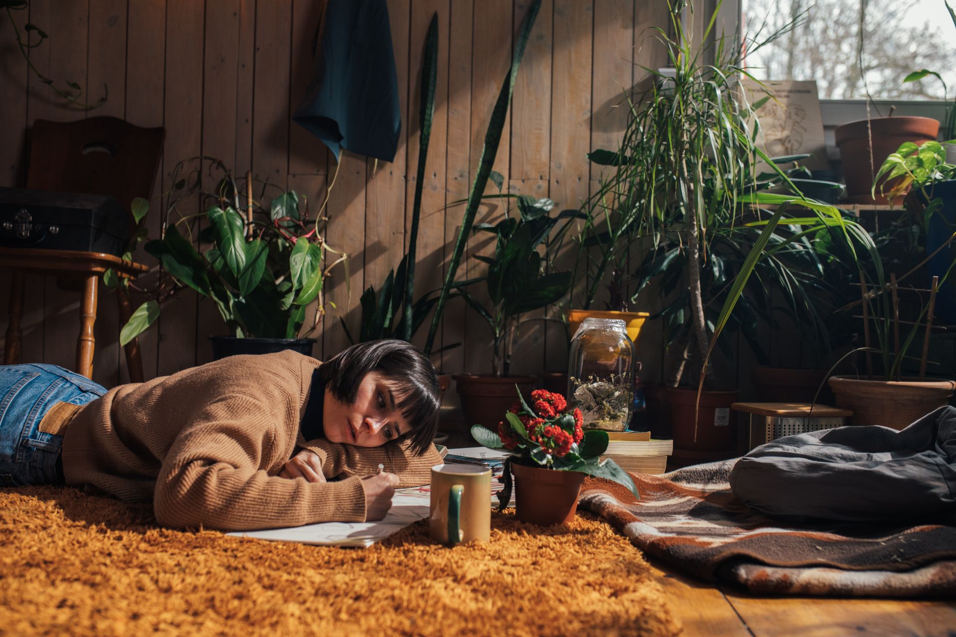 Adult woman drawing on a carpeted floor surrounded by indoor plants in cozy natural light.