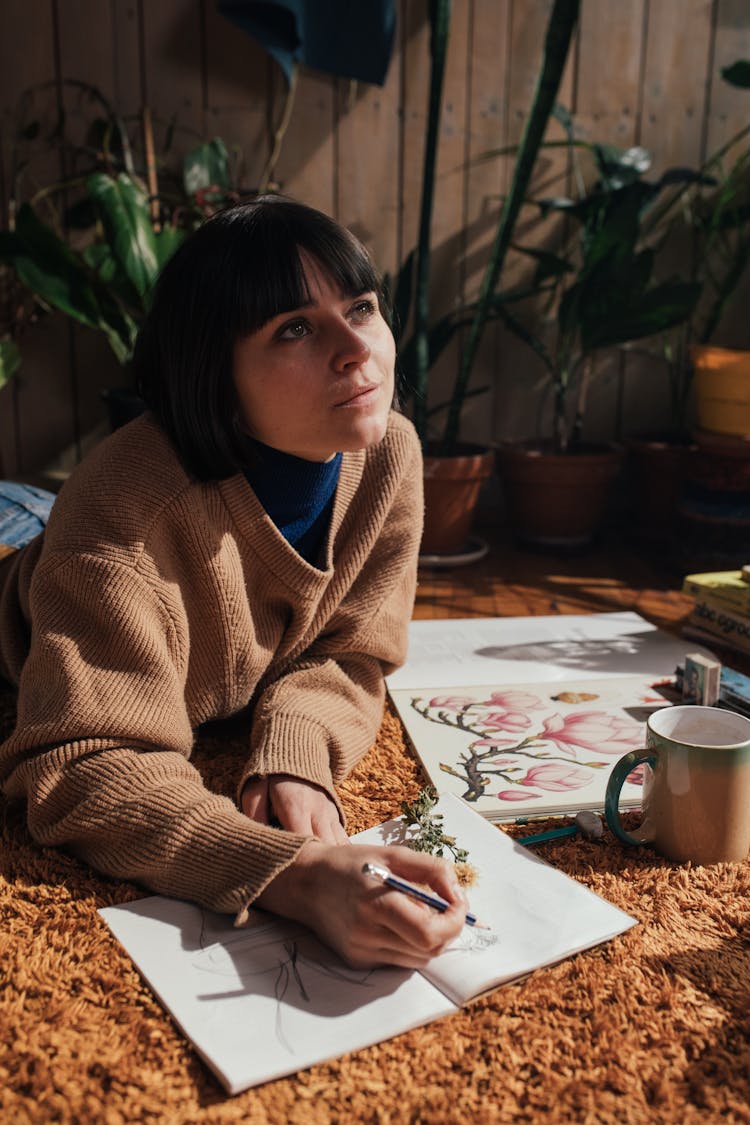 A Woman In A Sweater Lying Down On A Carpet While Drawing