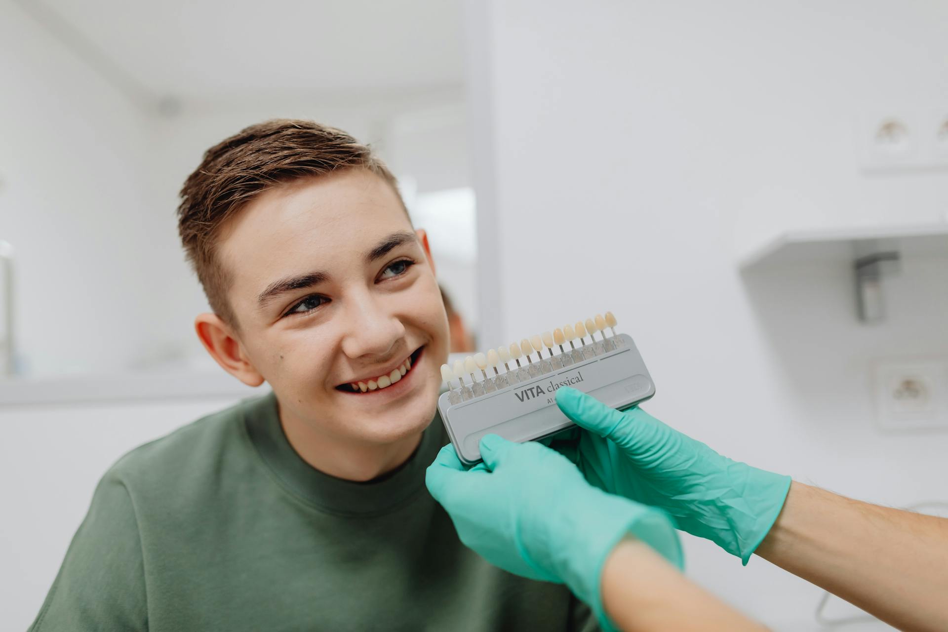 A Boy Smiling while a Person Wearing Latex Gloves is Holding a Dental Shade Guide