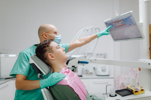 Dentist in green scrubs shows a patient an X-ray on a monitor in a modern office setting.