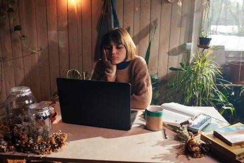 Free A woman in a cozy room surrounded by plants, appearing bored while using her laptop. Stock Photo
