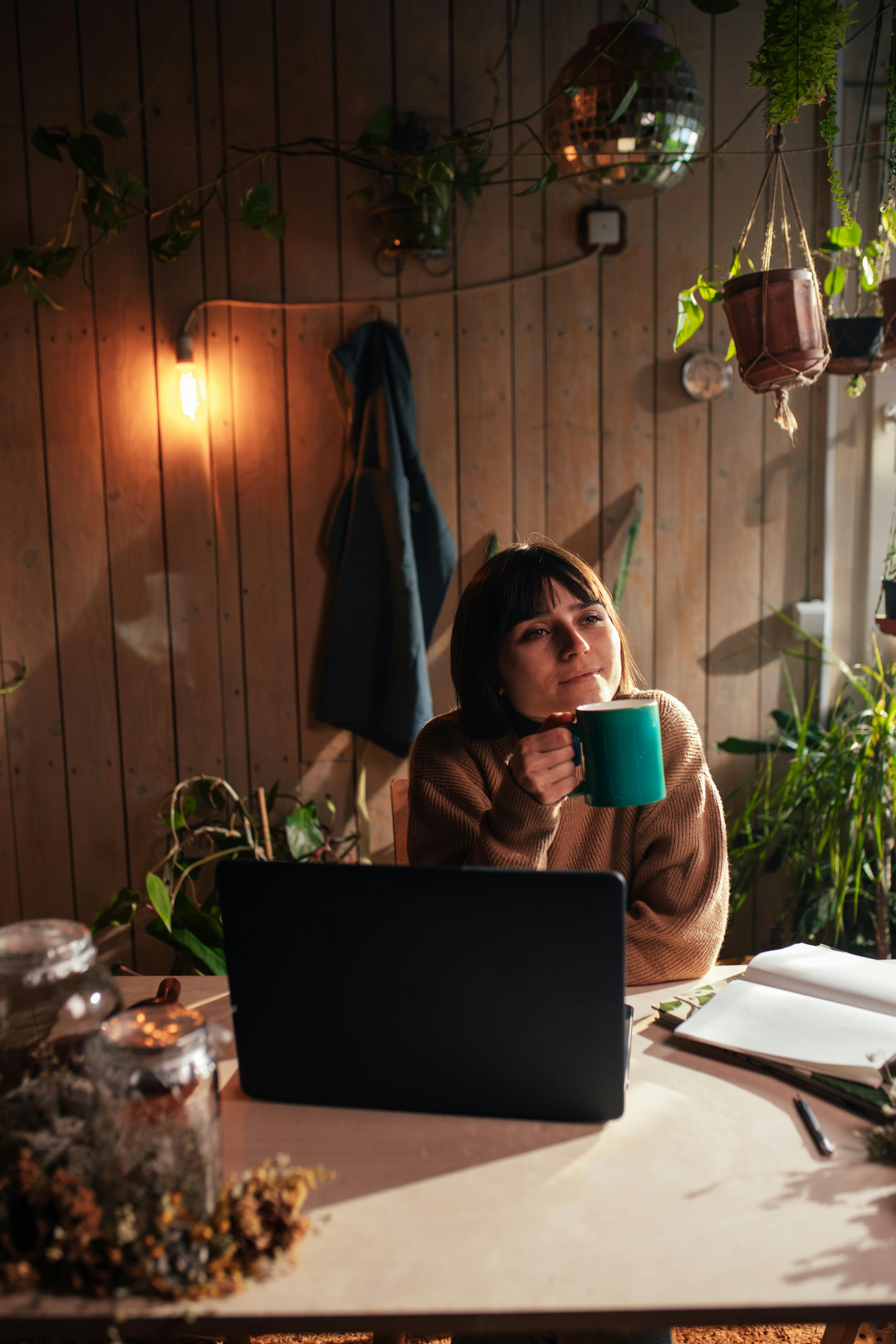 A woman enjoys a drink in a cozy indoor workspace with houseplants and warm lighting.