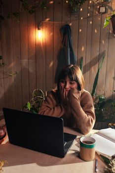 Woman in a knitted sweater working in a cozy, plant-filled room with laptop and coffee.