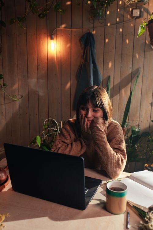 Free Woman in a knitted sweater working in a cozy, plant-filled room with laptop and coffee. Stock Photo