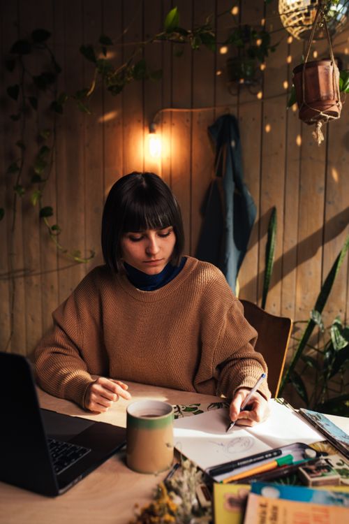 Free Woman drawing at a cozy study table with a coffee mug and laptop, surrounded by plants. Stock Photo