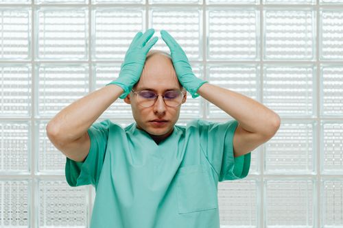 Free A healthcare worker in green scrubs looking stressed, hands on head with eyeglasses. Stock Photo