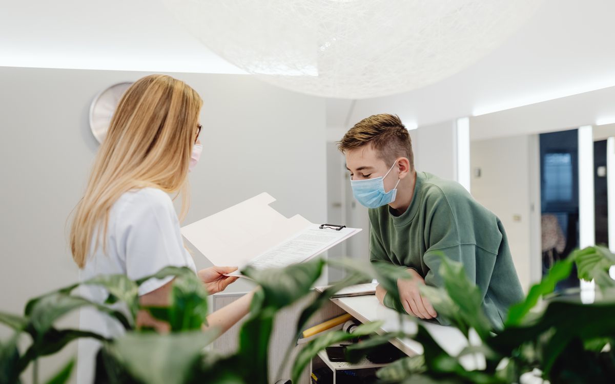 A woman and man in face masks converse over documents at a clinic counter indoors.