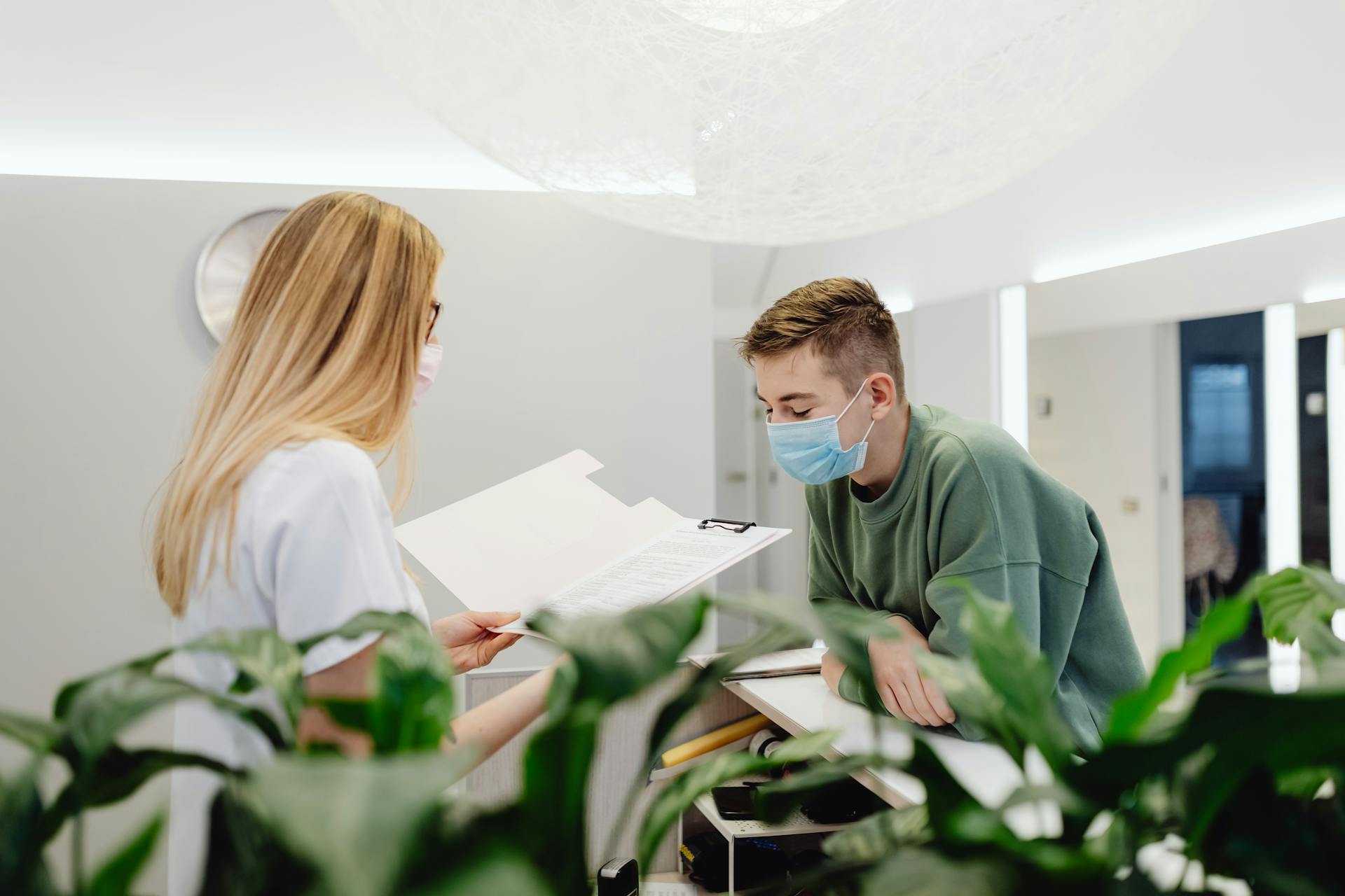 A woman and man in face masks converse over documents at a clinic counter indoors.