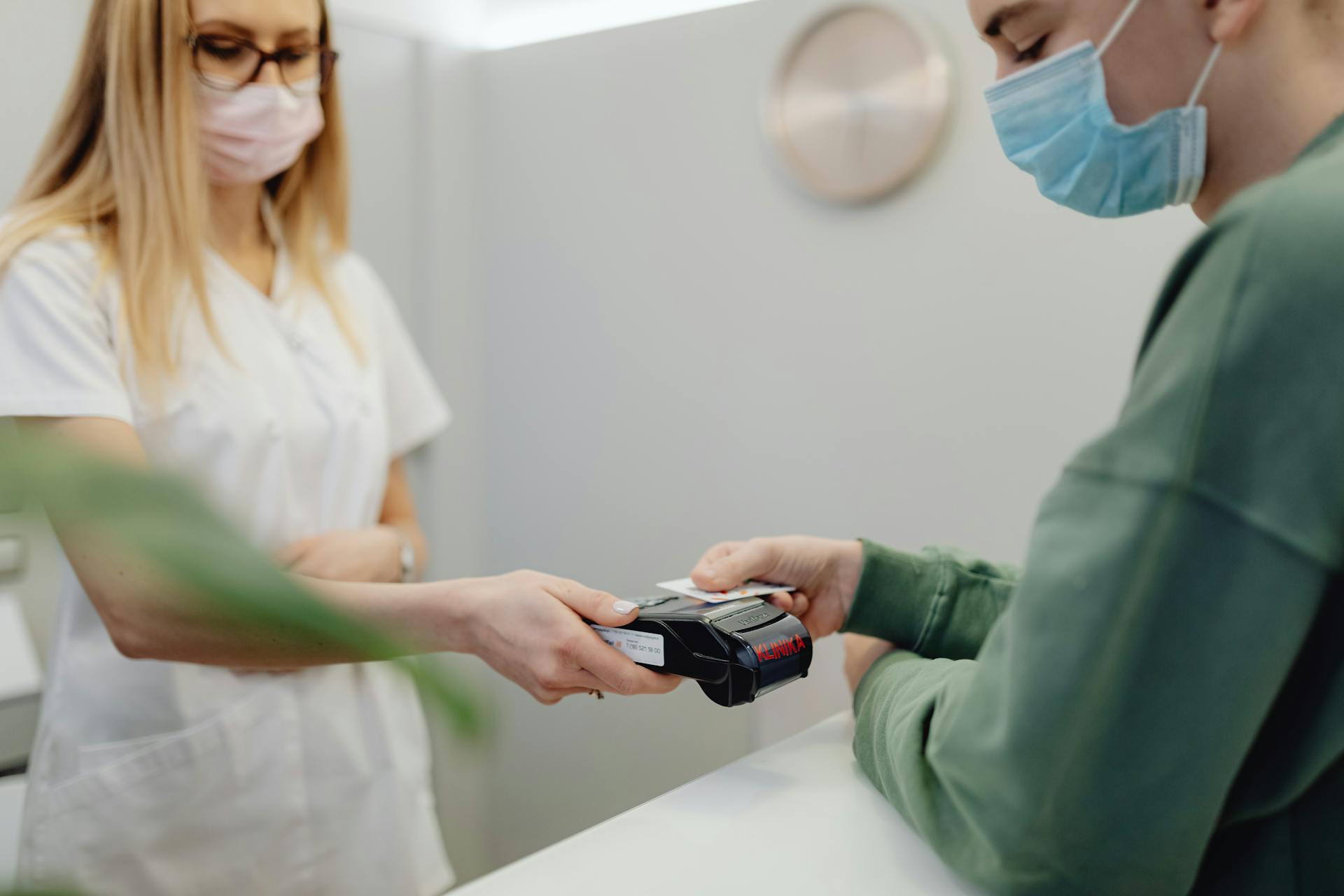 Person making a payment with a credit card at a medical reception desk, both parties wearing masks.