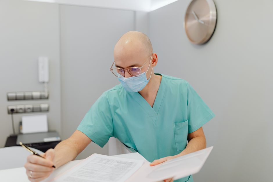 Trabajador de la salud masculino con uniformes médicos y mascarilla escribiendo notas en una oficina médica.