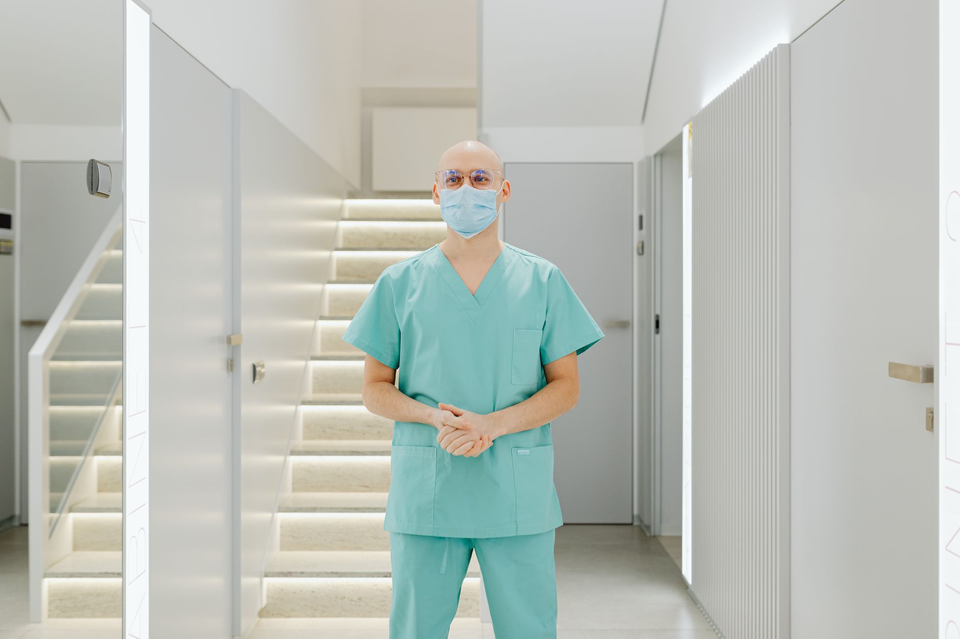 Healthcare worker in scrub suit standing indoors with face mask in clinic hallway.
