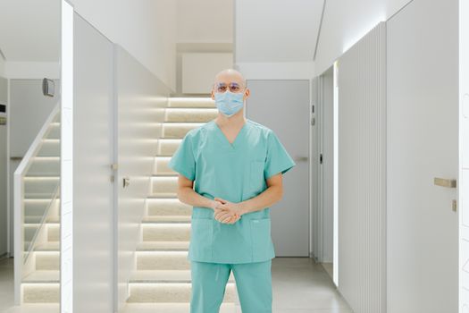 Healthcare worker in scrub suit standing indoors with face mask in clinic hallway.
