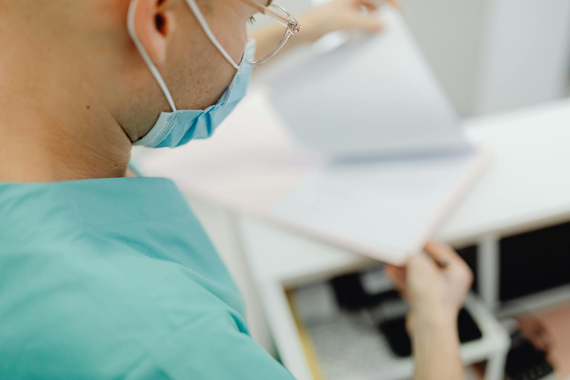 A medical professional in scrubs and mask examines documents in a clinical setting.