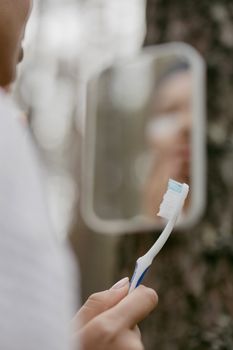 Person brushing teeth outdoors in front of a mirror with natural blurred background.