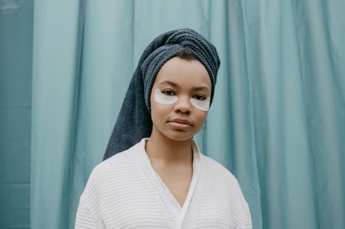 Free Woman in a white robe wearing eye patches, standing indoors with a head towel against a curtain backdrop. Stock Photo