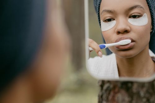 Free A black woman in a bathroom brushes her teeth while wearing cosmetic eye patches in front of a mirror. Stock Photo