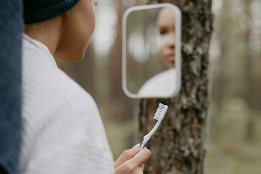 A person holding a toothbrush reflected in a mirror attached to a tree outdoors.