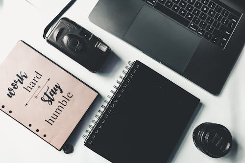 Flat lay of modern black laptop with film photo camera and notebooks placed on white table
