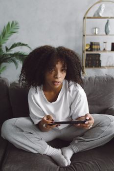 Young woman with afro hair playing video games indoors, sitting on couch in casual attire.