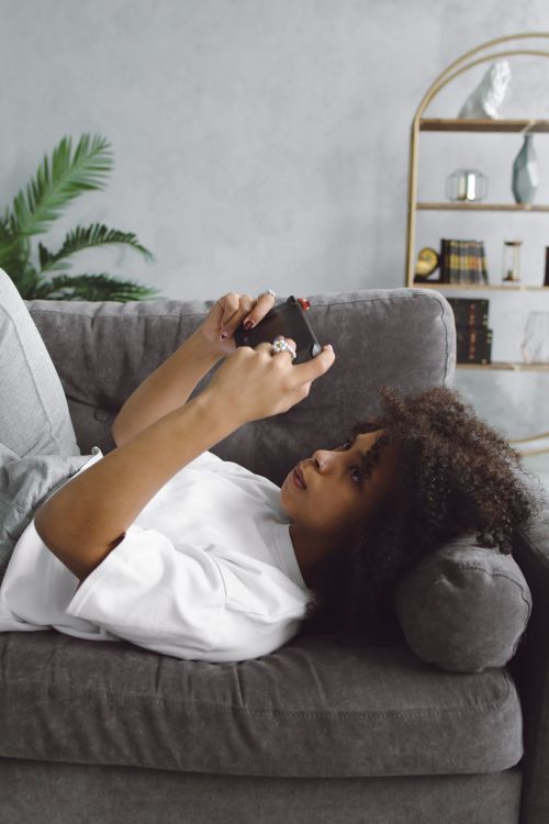 Free African American woman lying on a couch playing video games, capturing a cozy and casual indoor moment. Stock Photo