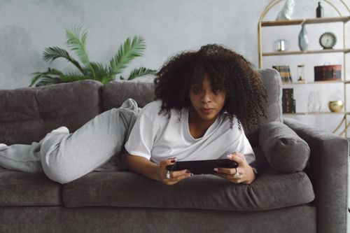 Free A woman with curly hair wearing gray pants, lying on a couch, holding a digital tablet. Stock Photo