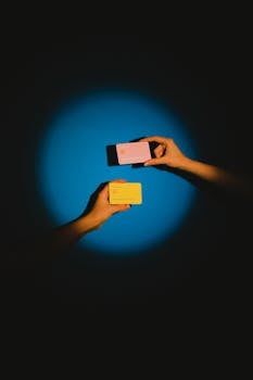Two hands exchanging colorful credit cards under a spotlight against a vibrant blue background, symbolizing modern financial transactions.