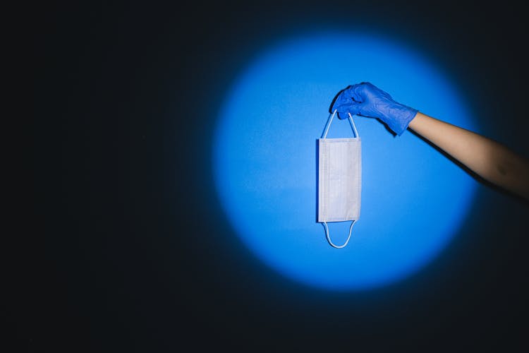 Hand In Blue Surgical Glove Holding A White Face Mask In Blue Spot Light