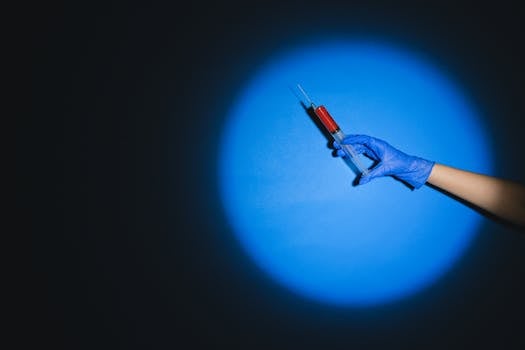 A gloved hand holds a syringe filled with red liquid against a dramatic blue spotlight.