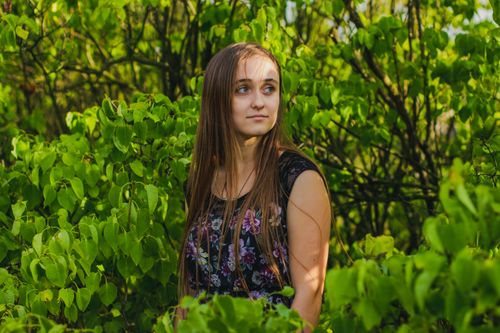 Free Portrait of a young woman in a floral dress amidst vibrant green leaves on a sunny day. Stock Photo