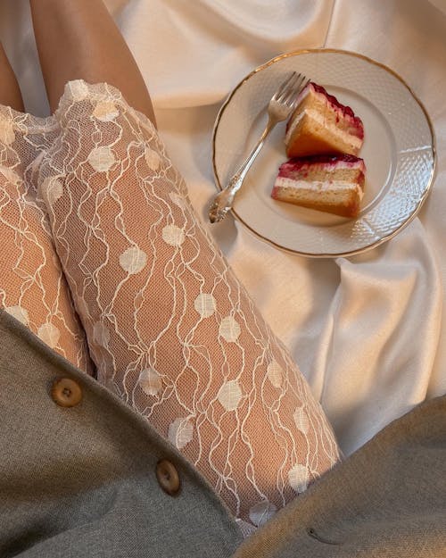 Top view of anonymous female wearing transparent cloth sitting on silk creased fabric with piece of cake on plate in room