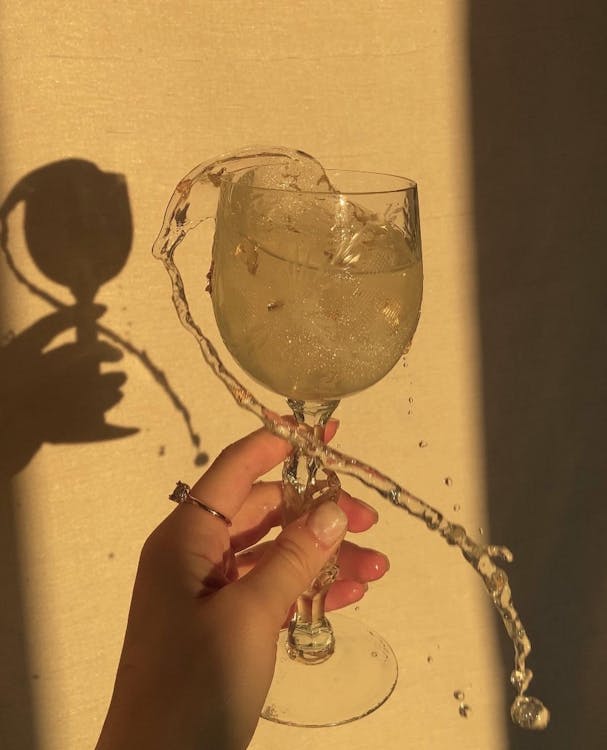 Unrecognizable female with transparent wineglass of splashing alcohol beverage standing near light wall with shadow in bright sunlight in room