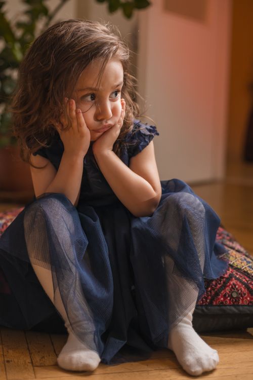 Free A young girl sits thoughtfully indoors in a blue dress, resting her face in her hands. Stock Photo
