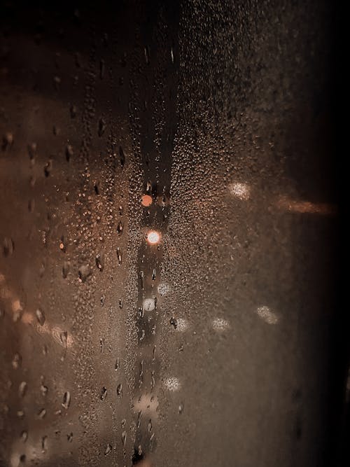 Abstract blur of street lights of city under dark sky reflecting through wet glass of window with droplets of rain in foggy weather at night
