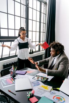 Two colleagues engaged in a discussion in a modern office setting with documents and laptops visible.