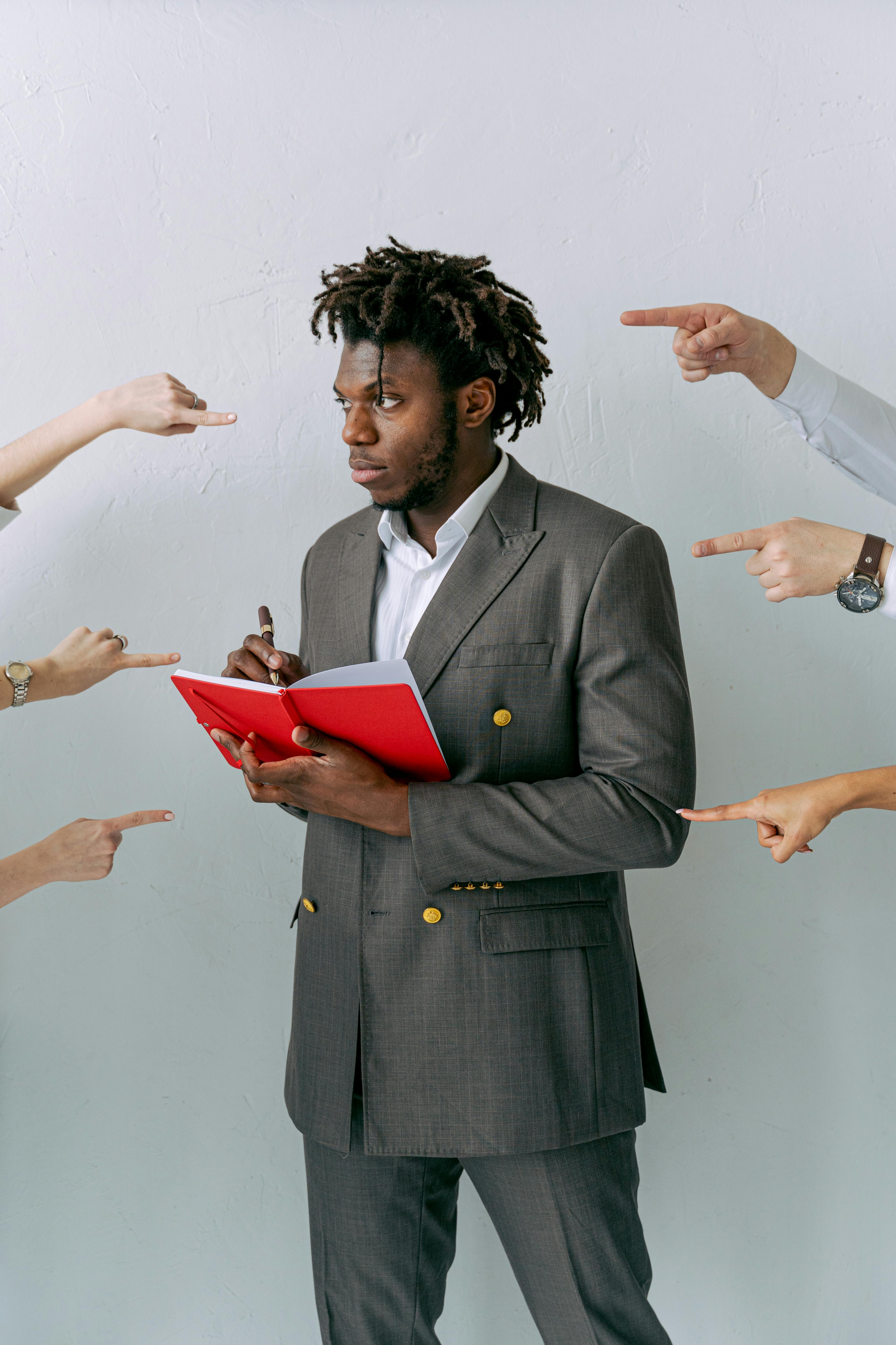 Businessman holding a red notebook surrounded by pointing hands. Concept of pressure and decision-making.